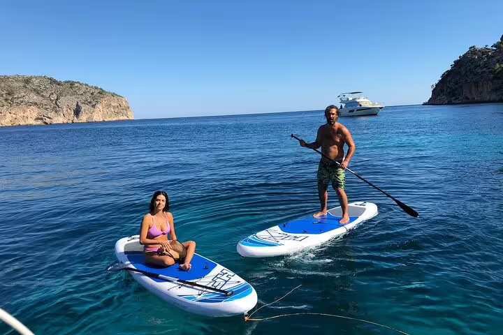 Two people paddleboarding in crystal-clear waters near a catamaran on a sunny Formentera and Ibiza group tour.