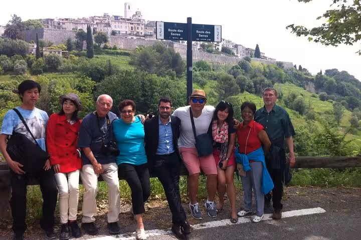 Group of tourists posing in front of a scenic hillside village on a guided tour of the French Riviera from Nice.