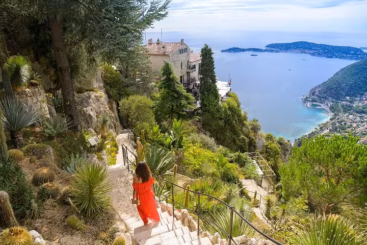Woman in red dress descending stone steps with a panoramic view of the French Riviera coastline near Nice.