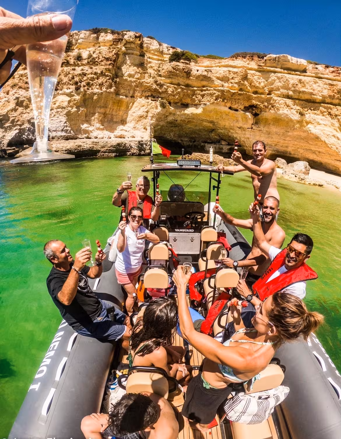 Friends raising champagne glasses on a tailor‑made boat charter, cruising turquoise Algarve caves under a bright summer sky