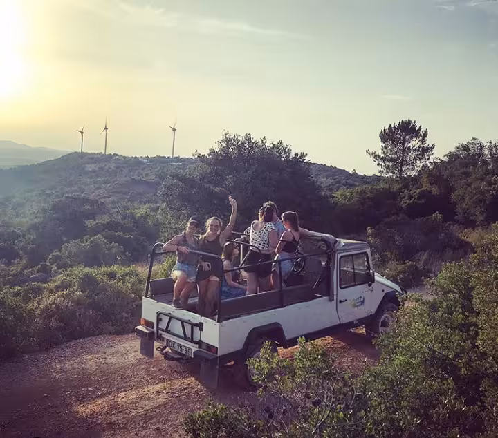 Friends on open-top jeep at golden hour, enjoying sunset sparkling wine tour with wind turbines on distant hills