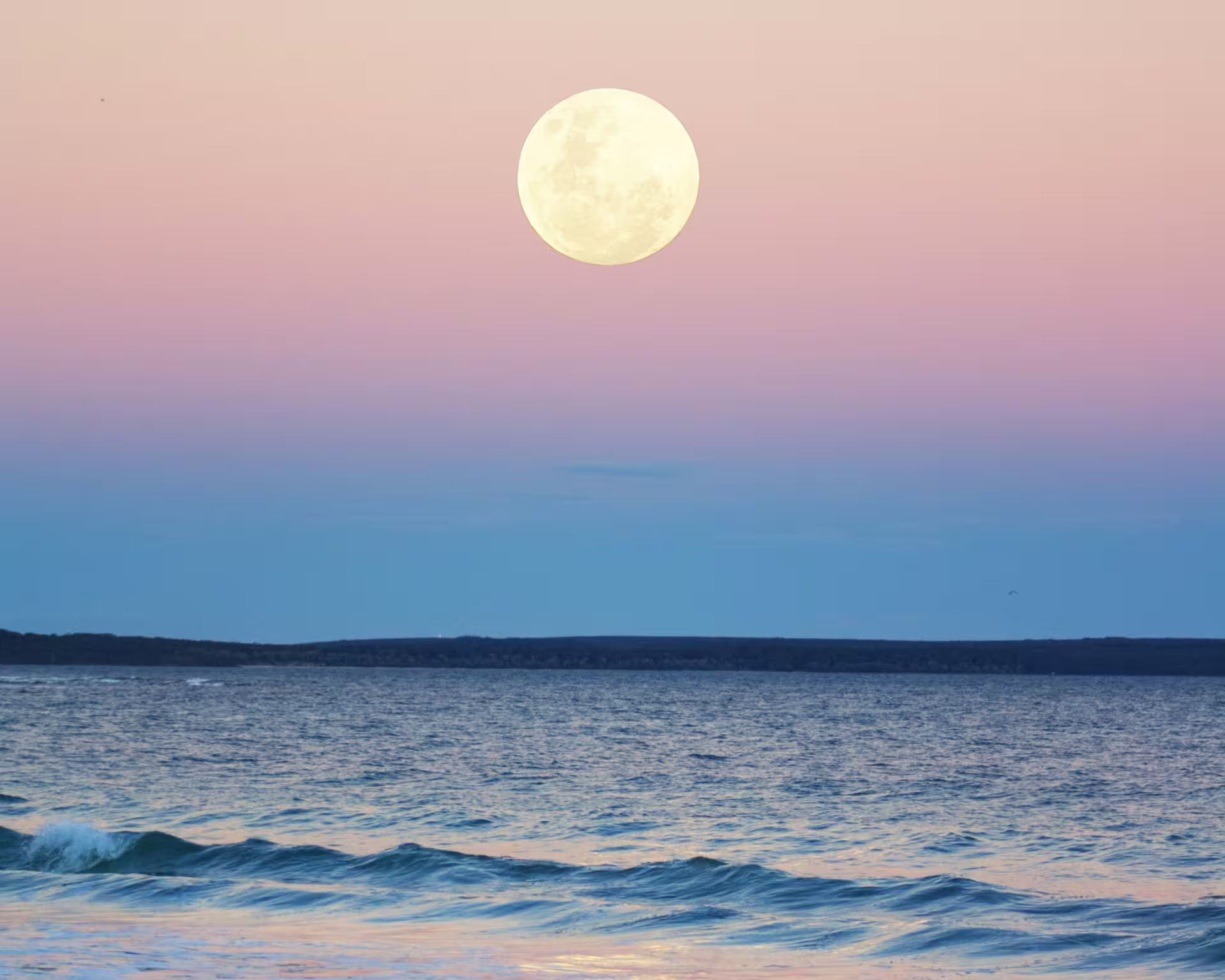 Full Moon even with light, sky degraded in shades of blue, purple, gold, pink, yellow. Calm sea.