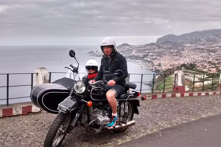 Two people on a motorcycle with sidecar enjoy a scenic view of Funchal Old Town and coastline, ideal for tour exploration.