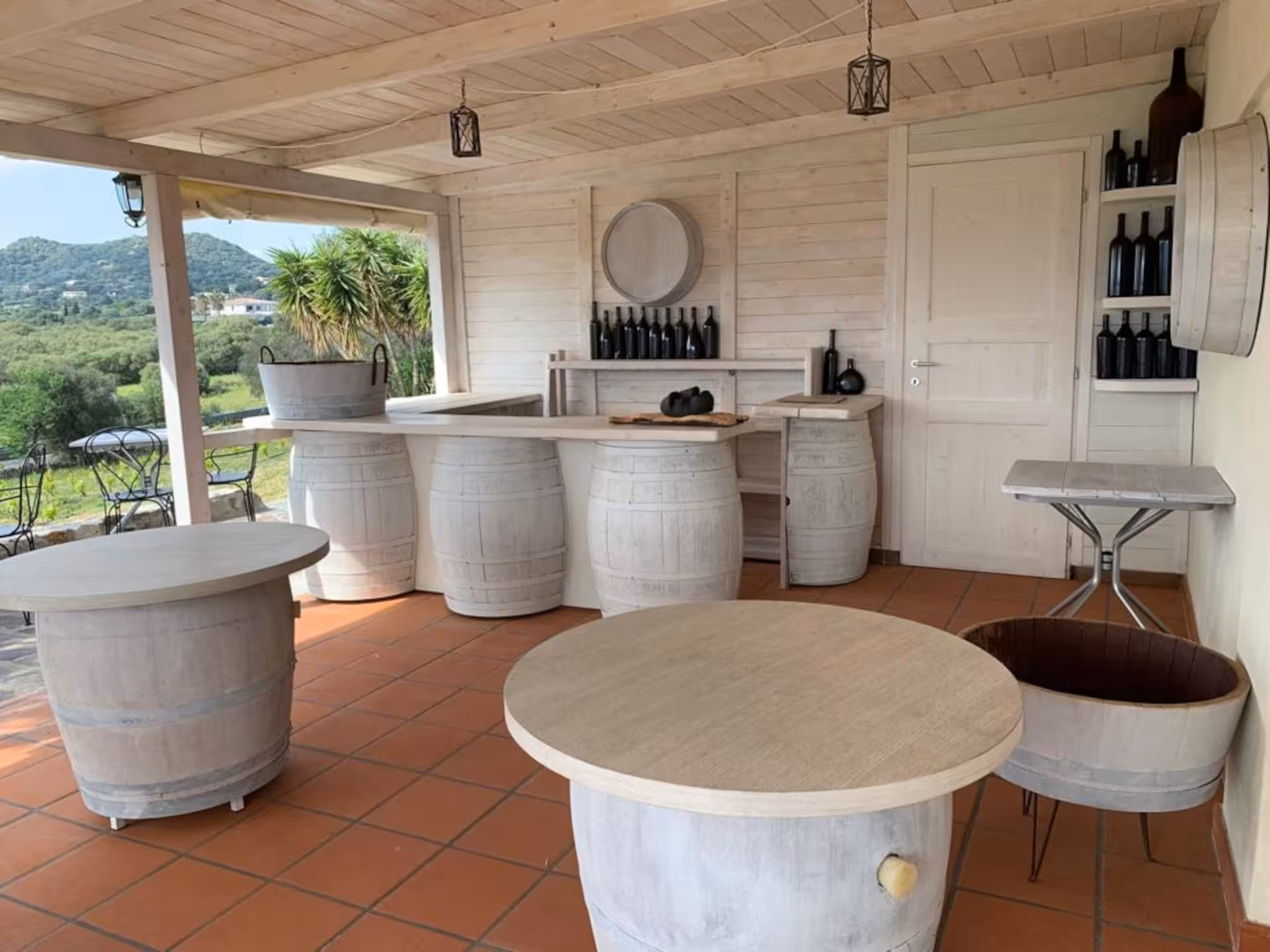 Rustic outdoor tasting area with barrel tables at a Gallura winery in Olbia, overlooking lush vineyards.