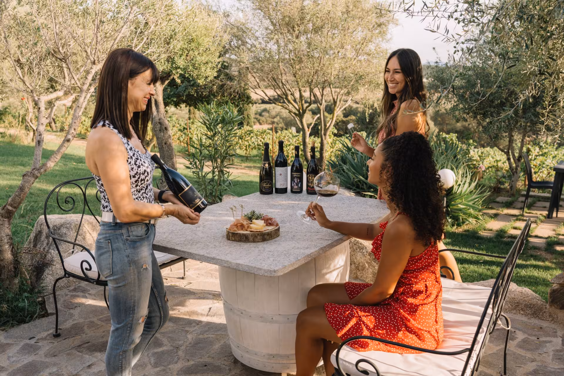 Women enjoying wine tasting at an outdoor table in a picturesque Gallura vineyard, Olbia, surrounded by lush greenery.