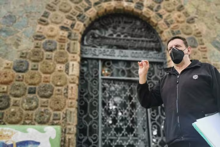 Guide in mask explains architectural details at Colonia Güell entrance, showcasing Gaudí's design on a skip-the-line private tour.