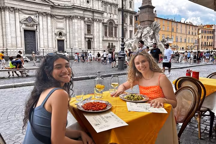 Two women enjoy gelato and pasta at an outdoor cafe in Piazza Navona, Rome, with a scenic backdrop.