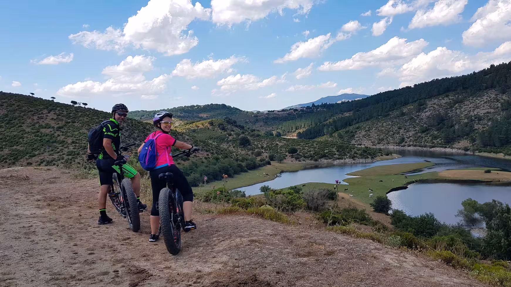 Cyclists on an ebike tour enjoy scenic views of Gennargentu's rolling hills and serene lake under a blue sky.