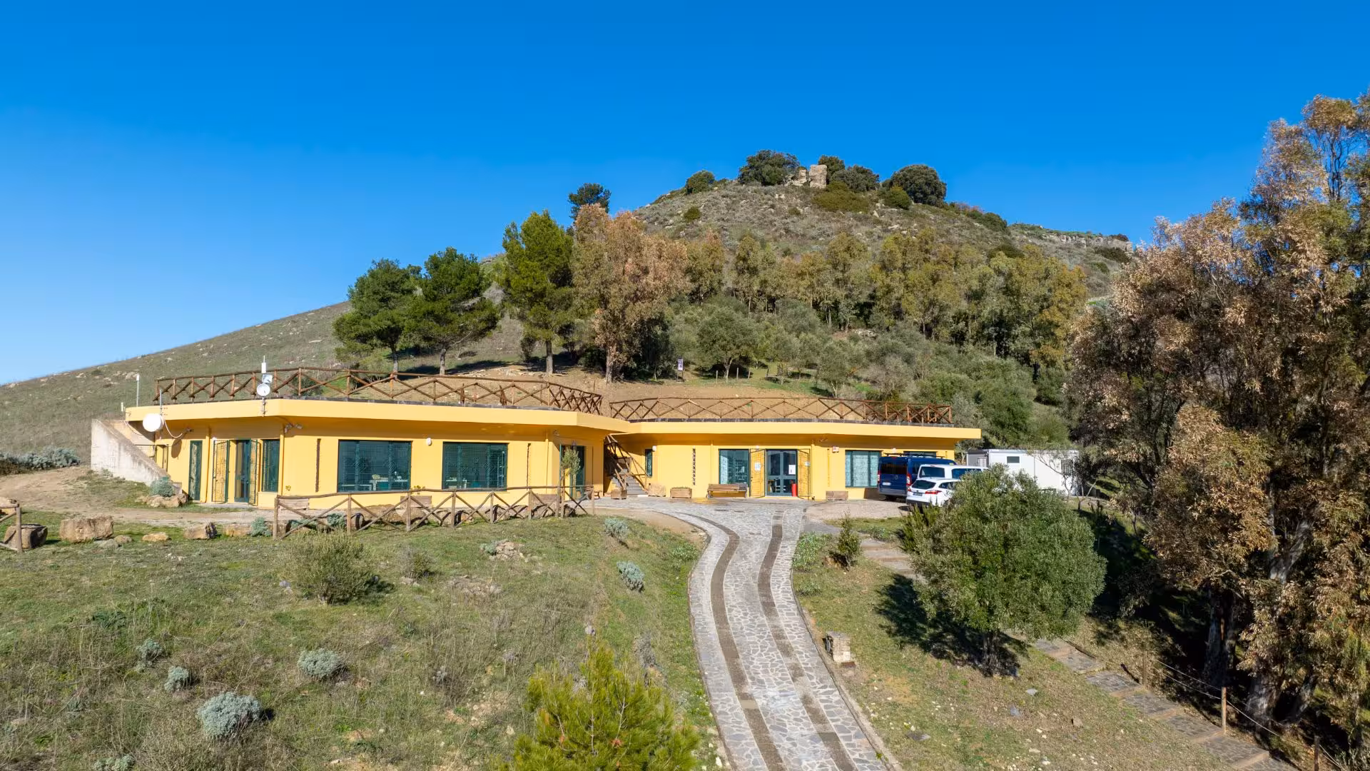 Charming yellow building at the foot of a hill in Giara Park, surrounded by lush greenery and vibrant blue skies.