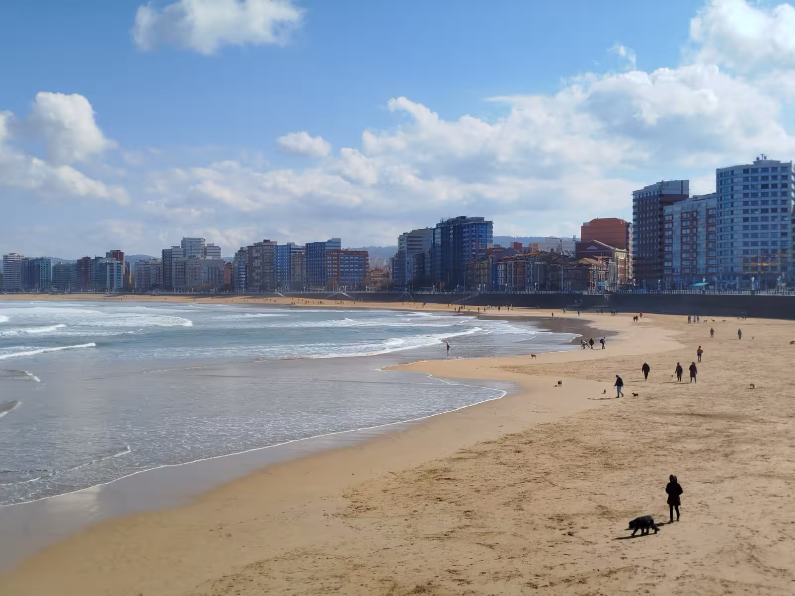 San Lorenzo Beach in Gijon with people strolling along the sandy shore, framed by the city skyline and blue skies.