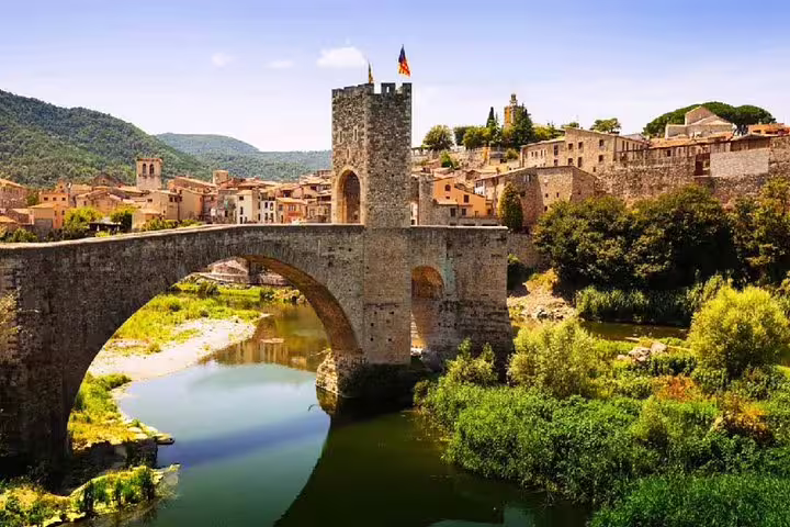 Medieval stone bridge and tower over a serene river in a Catalonian village, surrounded by lush greenery and historic architecture.