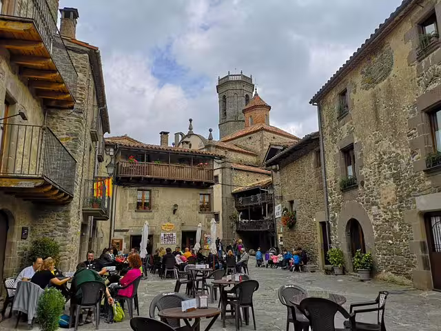 Visitors enjoy dining in a charming medieval village square in Catalonia, surrounded by historic stone buildings and a church tower.