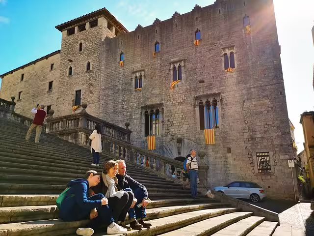 Tourists relax on sunlit stone steps in front of a medieval building in Catalonia, highlighting historic village charm.