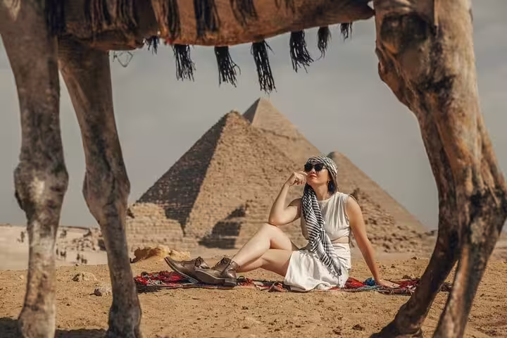 Traveler relaxing on desert rug framed by camel legs with Giza Pyramids on full-day tour with pickup