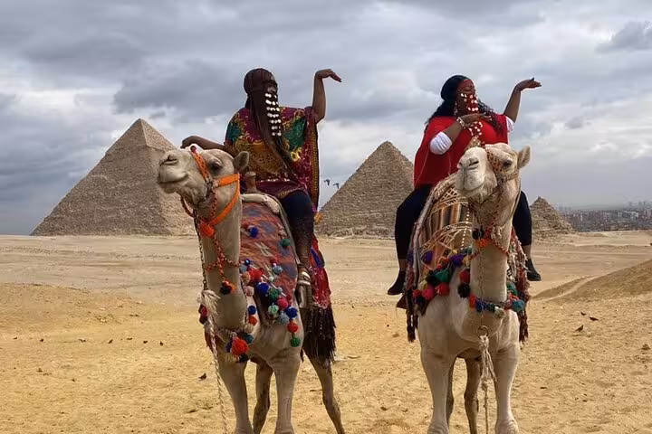 Two riders on decorated camels with Giza Pyramids behind, private camel ride tour with lunch in Cairo