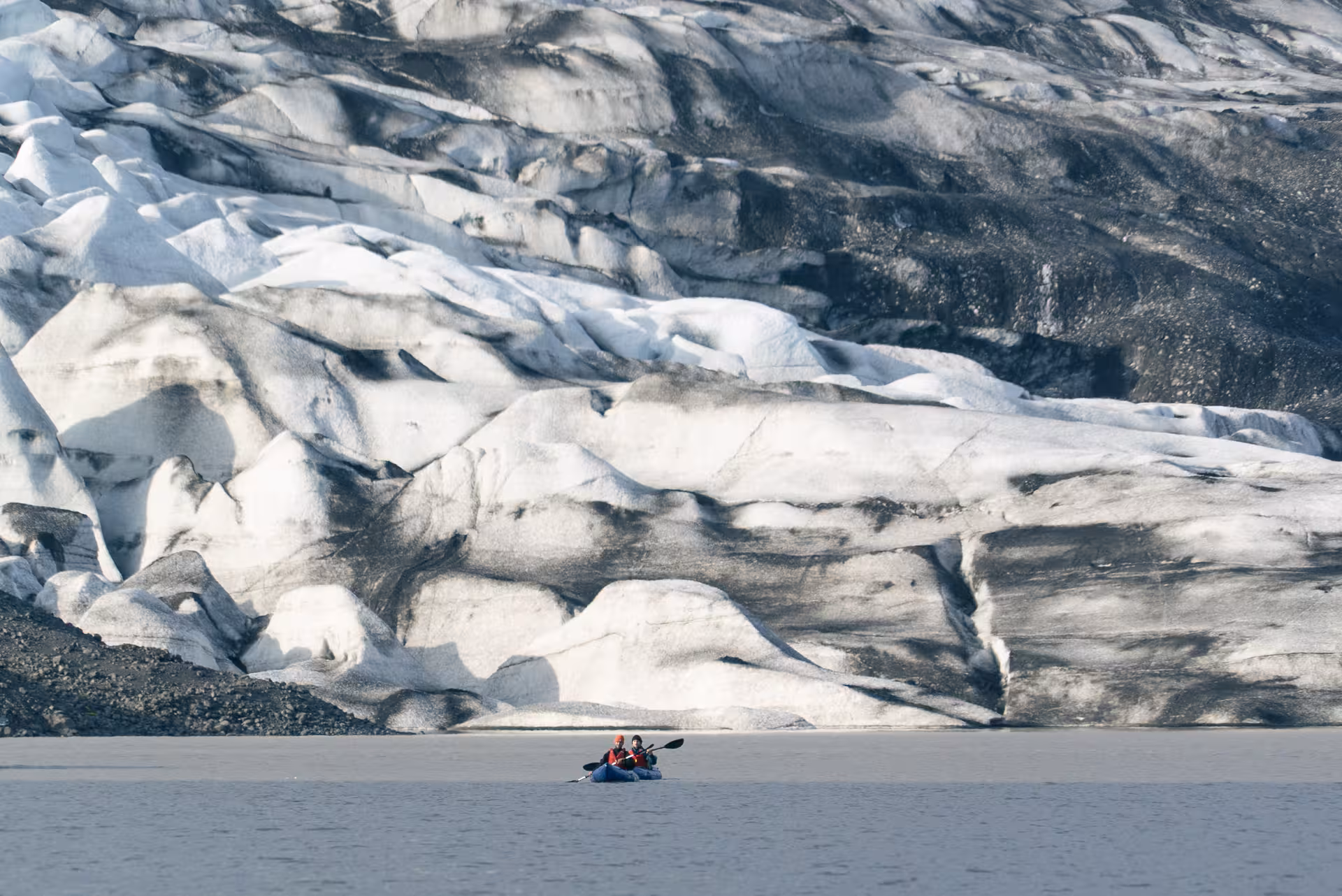 Two packrafters paddle a glacier lagoon beneath a towering ice wall on an Intro to Glacier Packrafting adventure