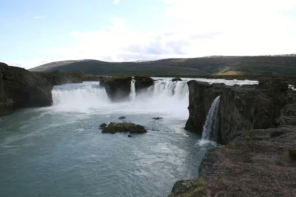 Godafoss waterfall cascading over rocky cliffs into turquoise waters with green hills in Iceland