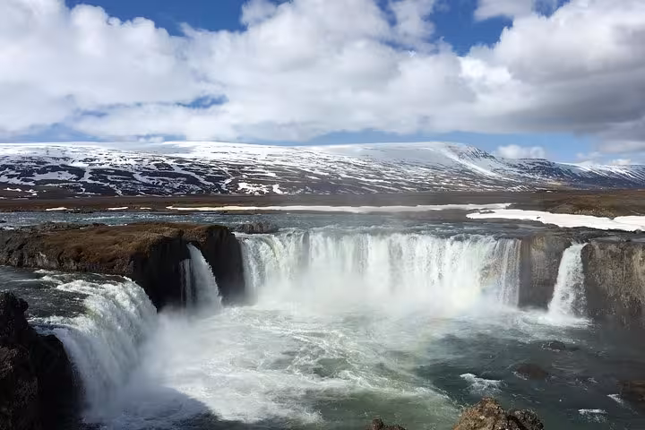 Majestic Godafoss waterfall cascading in Iceland's stunning landscape, featuring snow-capped mountains under a blue sky.