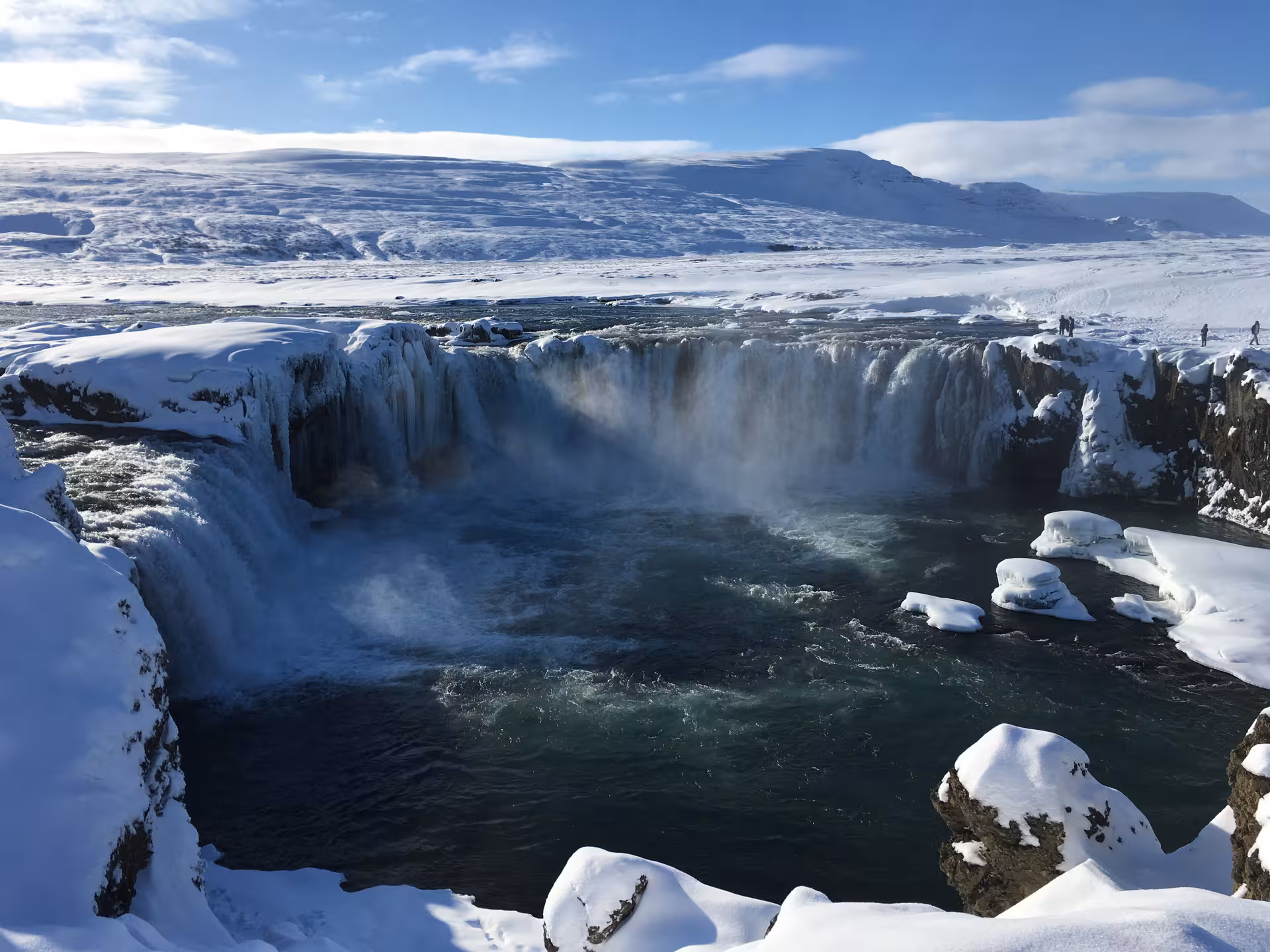 Majestic Godafoss Waterfall surrounded by snow and ice under a clear blue sky on a winter tour in Iceland.