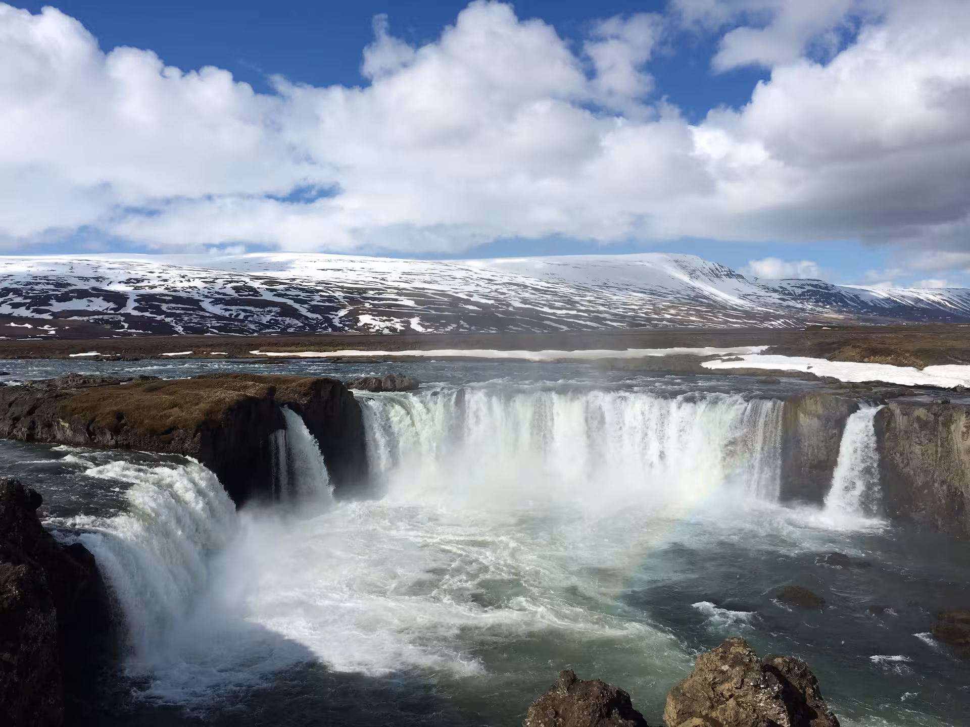 The majestic Godafoss waterfall cascades powerfully with a rainbow arching over the snowy landscape.