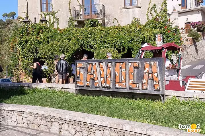 Savoca sign with tourists exploring a scenic stop on The Godfather tour in Sicily, featuring lush greenery and historic charm.