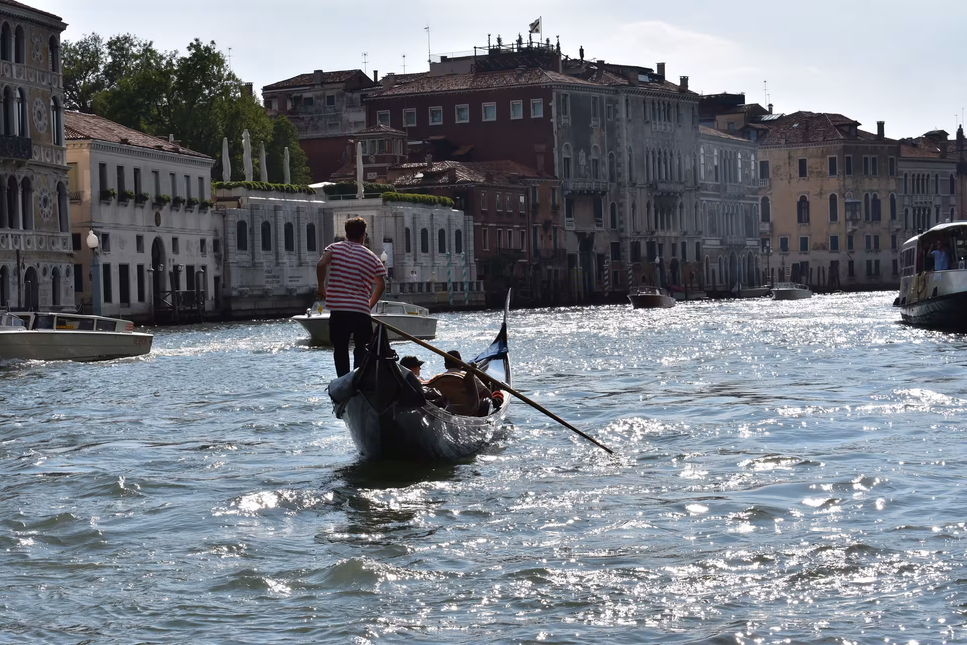 Gondolier steering a gondola down a sunlit Venice Grand Canal, lined with historic buildings.