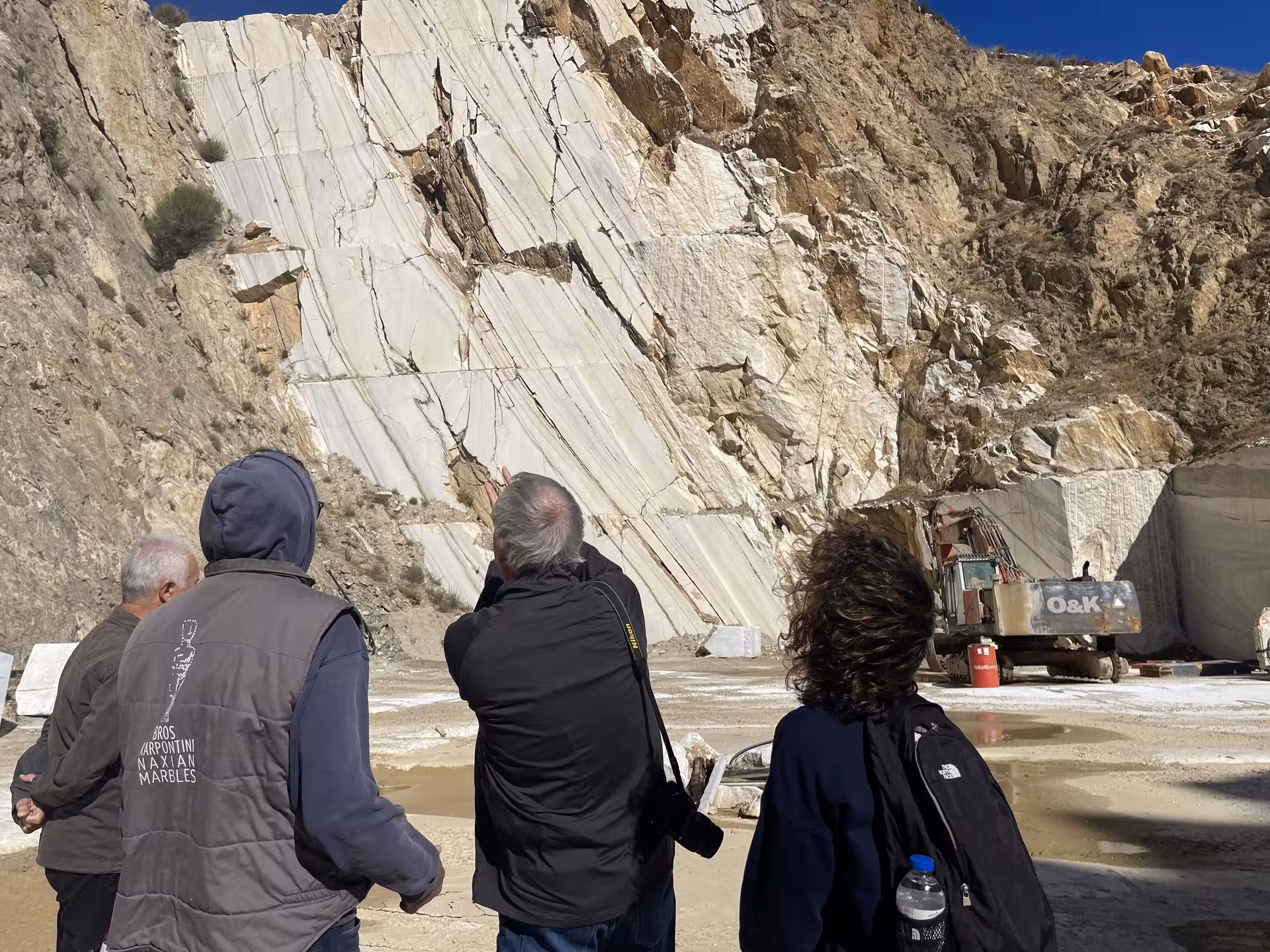 Visitors explore an active Greek marble quarry with towering white rock face and excavator on Marble World tour