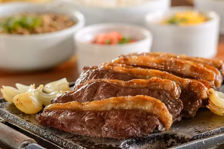 Grilled Brazilian steak served with sides, featured in Rio's Cookin' Good: 9-Dish & Caipis Fiesta.