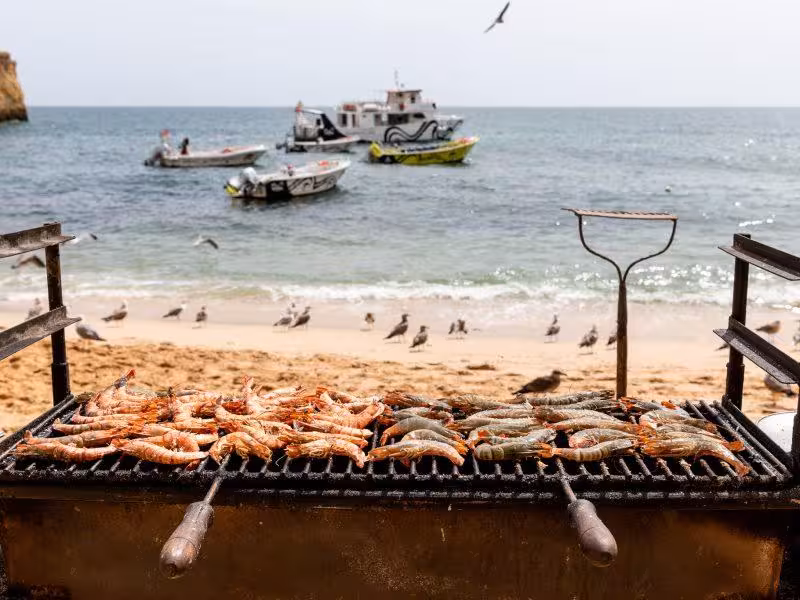 Grilled seafood sizzling on a beach barbecue with scenic ocean view and boats in the background.