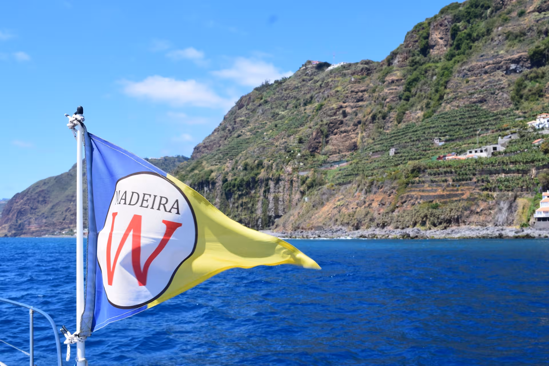 Madeira flag waving on GRINGO Yacht with lush coastal cliffs, ideal for private charter exploration.