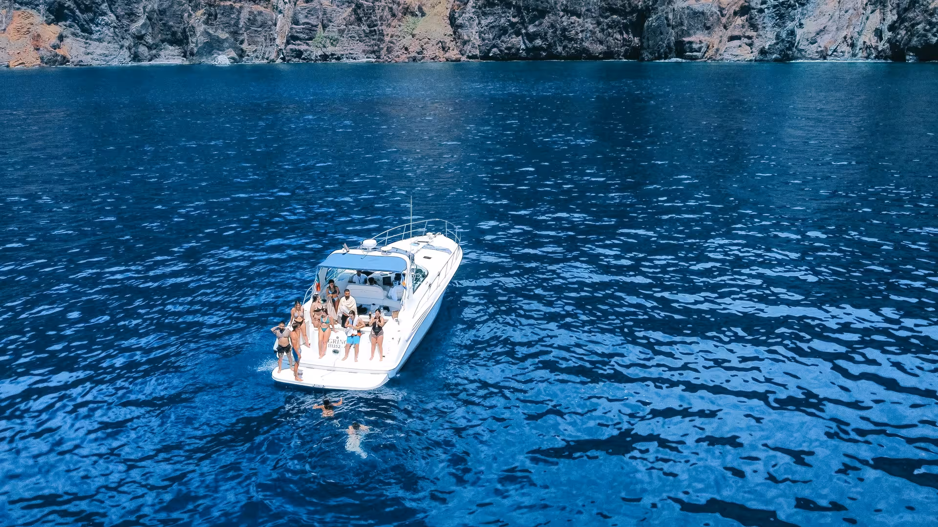 Aerial view of GRINGO Yacht with passengers enjoying a whale and dolphin watching cruise near rocky cliffs.