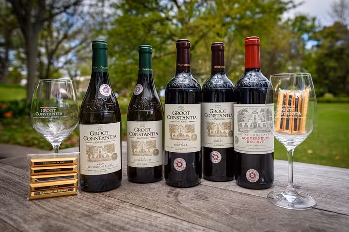 Selection of Groot Constantia wines elegantly displayed on a wooden table with branded wine glasses, set in a vibrant garden.