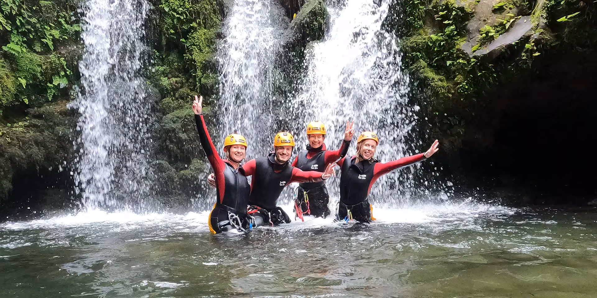 Group canyoning under a waterfall, wearing helmets and wetsuits, enjoying guided adventure canyon tour