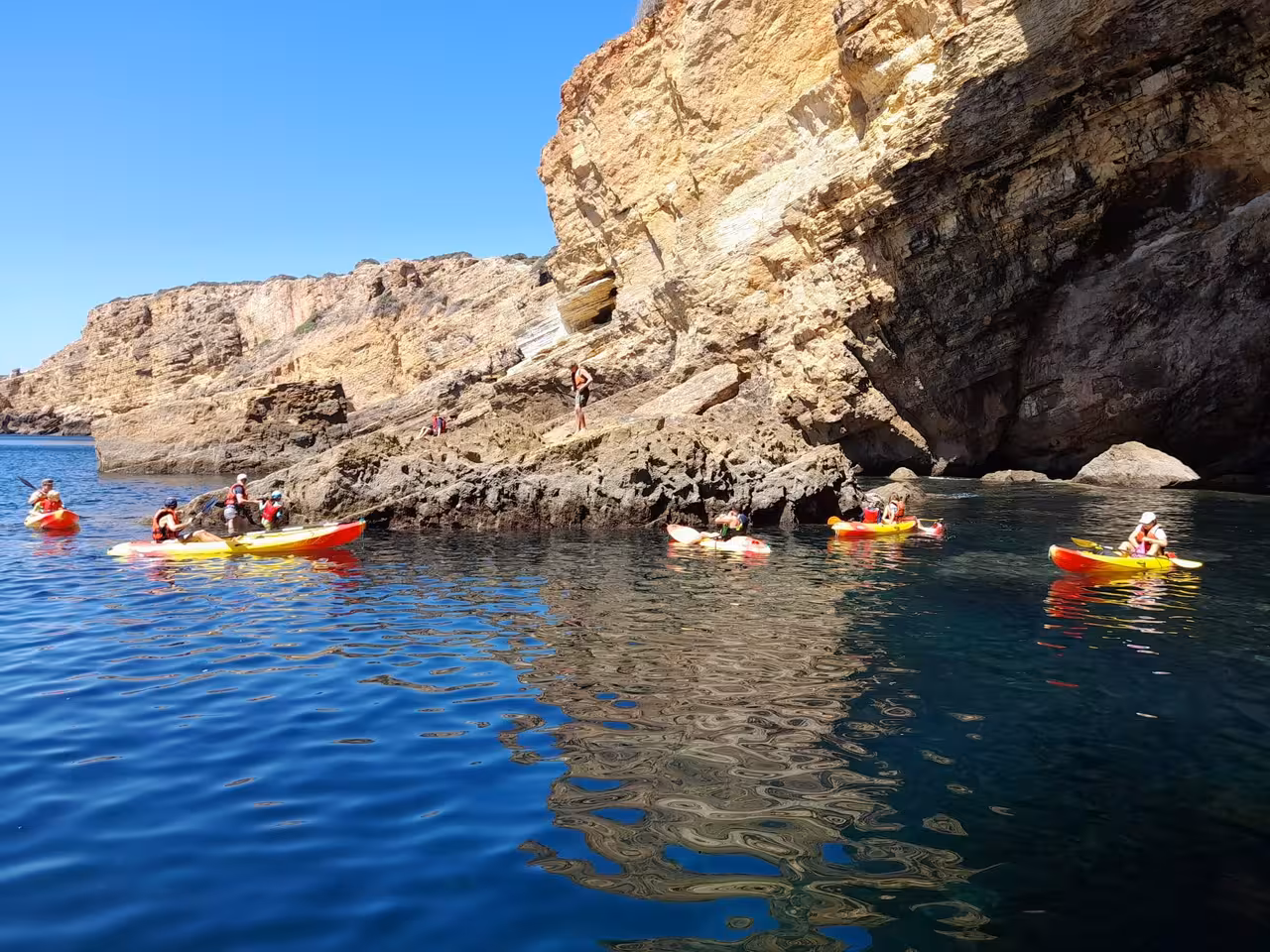 Group kayaking along rugged cliffs in clear blue waters on a sunny day, perfect for adventure seekers and nature lovers.