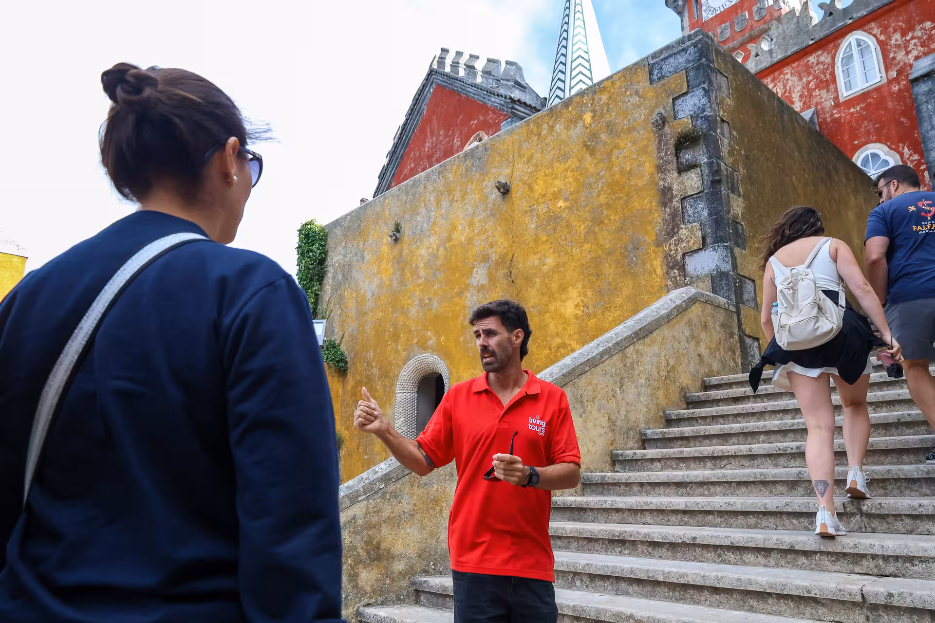 Guide in red shirt explaining to visitors on stone steps of the colorful Pena Palace in Sintra.