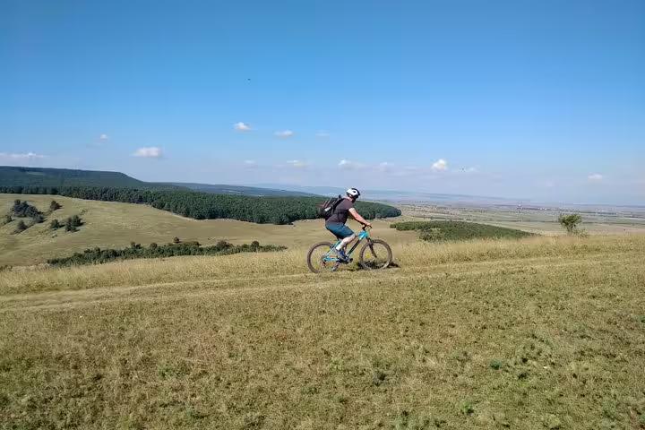 Cyclist riding a blue mountain bike across rolling grassy hills in Transylvania with forest and plains in background