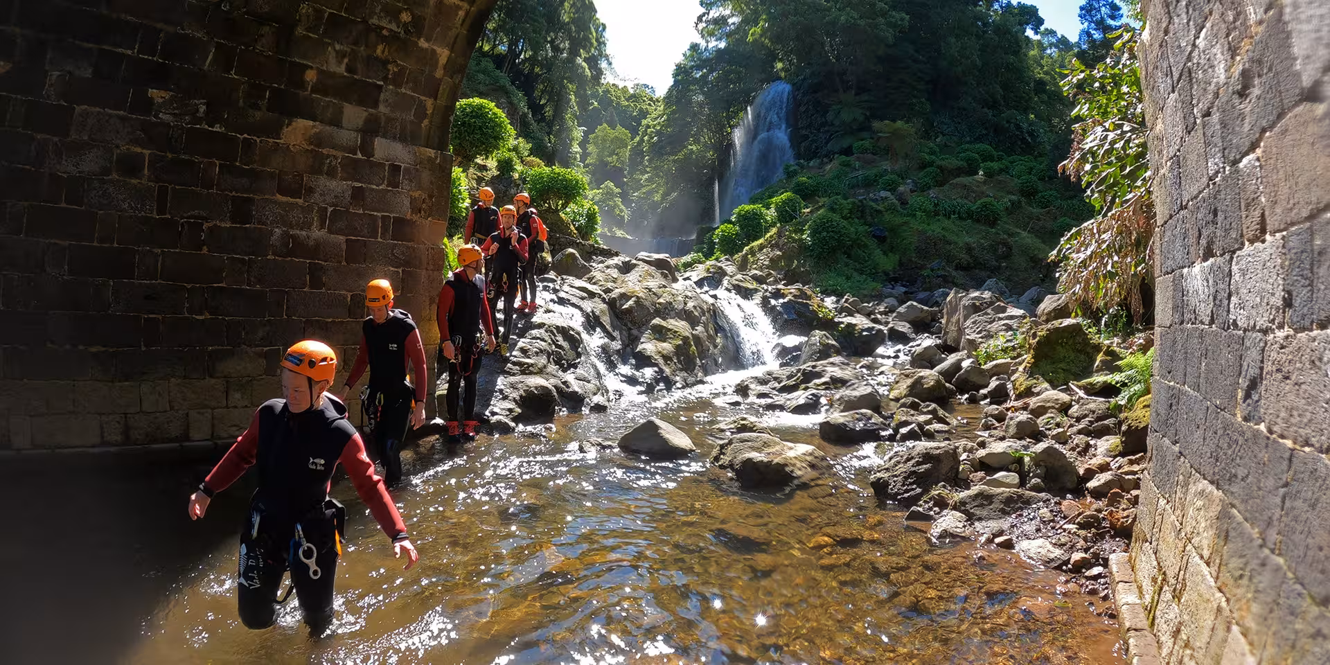 Guided canyoning group wading a rocky river under a stone bridge, heading toward a lush waterfall