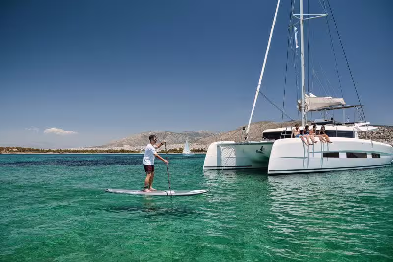 Friends relax on a modern catamaran while a guest paddleboards in clear turquoise water on a Gulf of Naples day tour