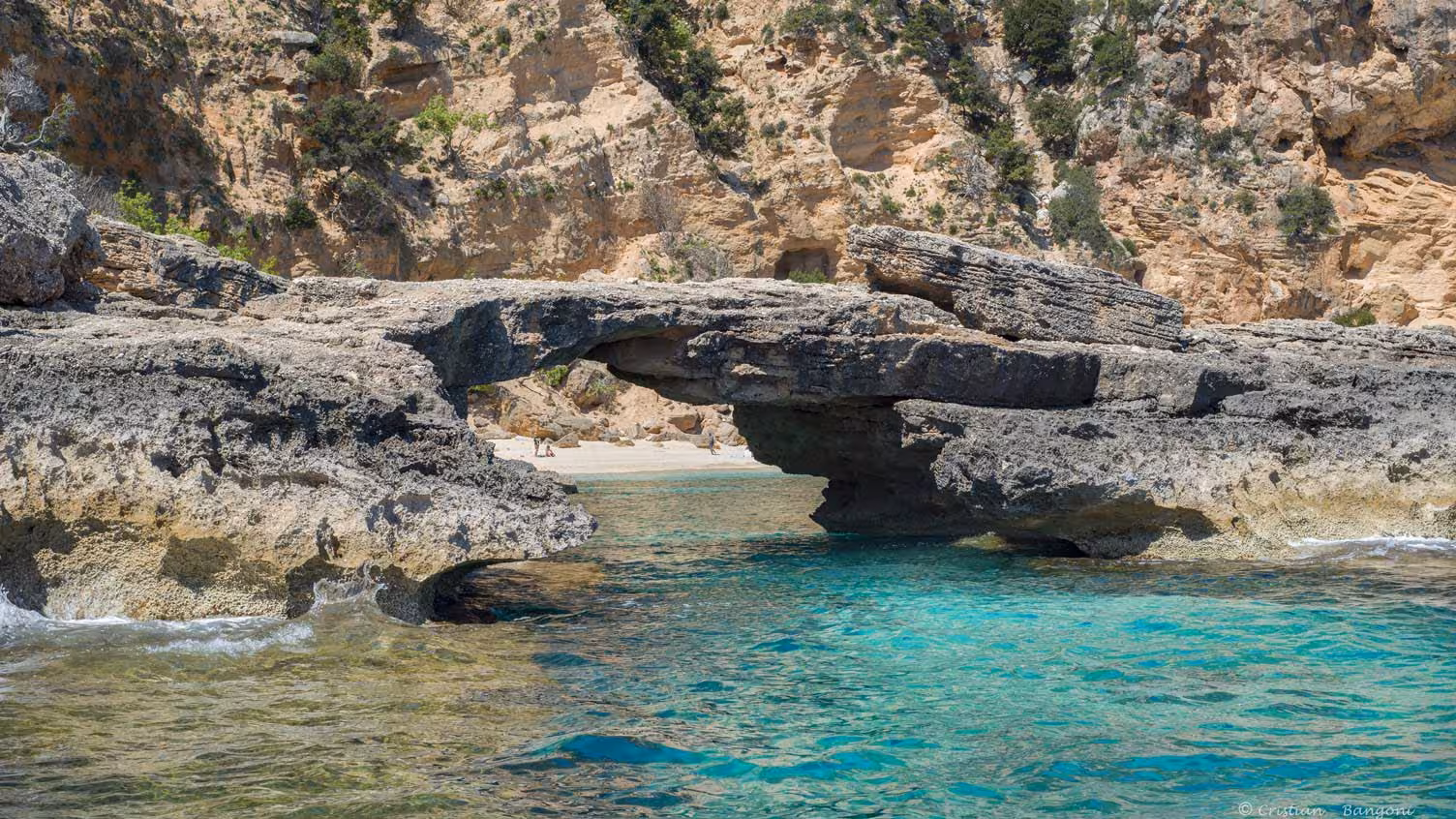 Natural rock arch over turquoise waters with rocky cliffs in the background in the Gulf of Orosei.