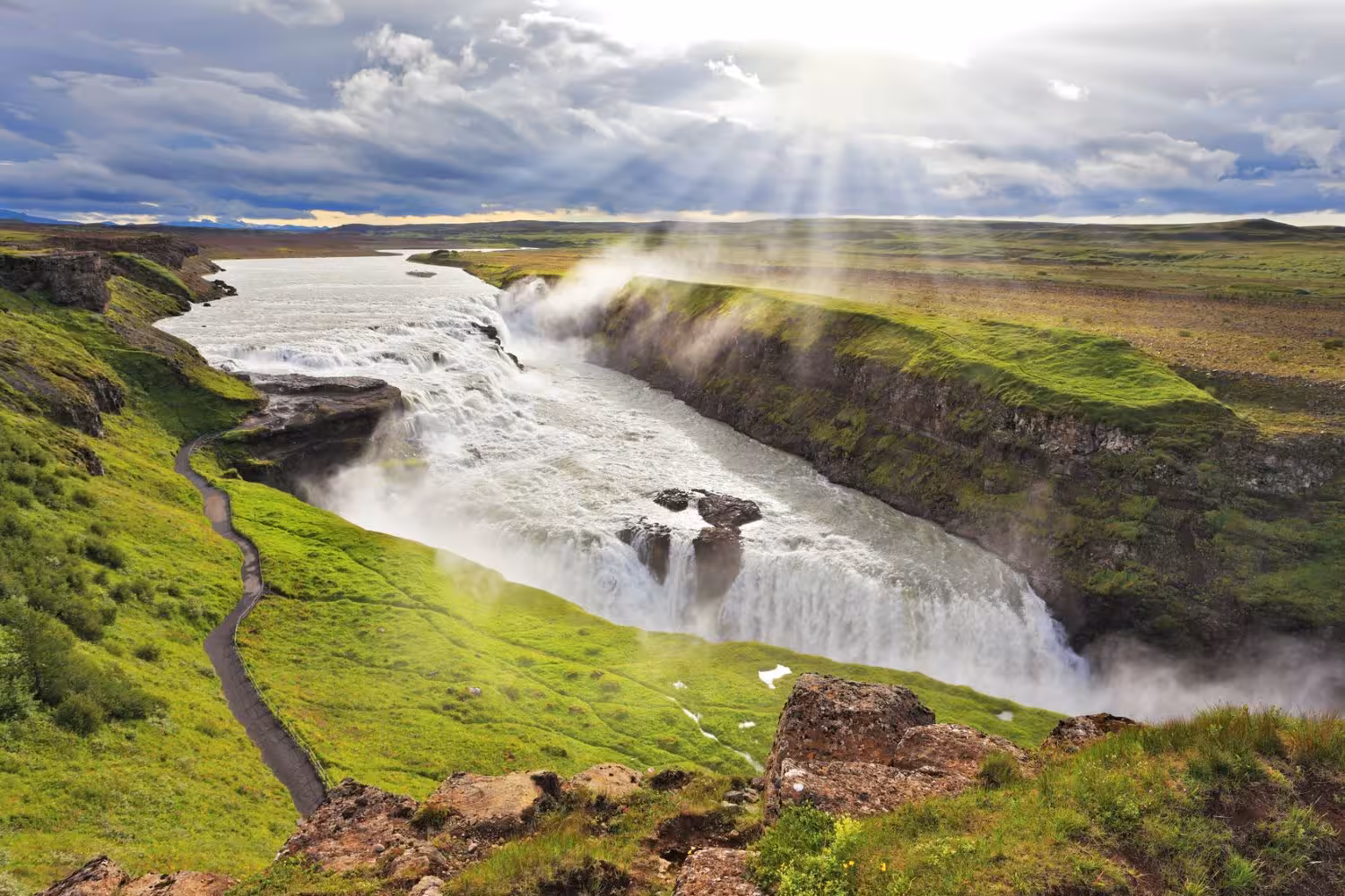 Sunlit Gullfoss waterfall in Iceland's Golden Circle cascades into a misty canyon surrounded by lush green landscape.