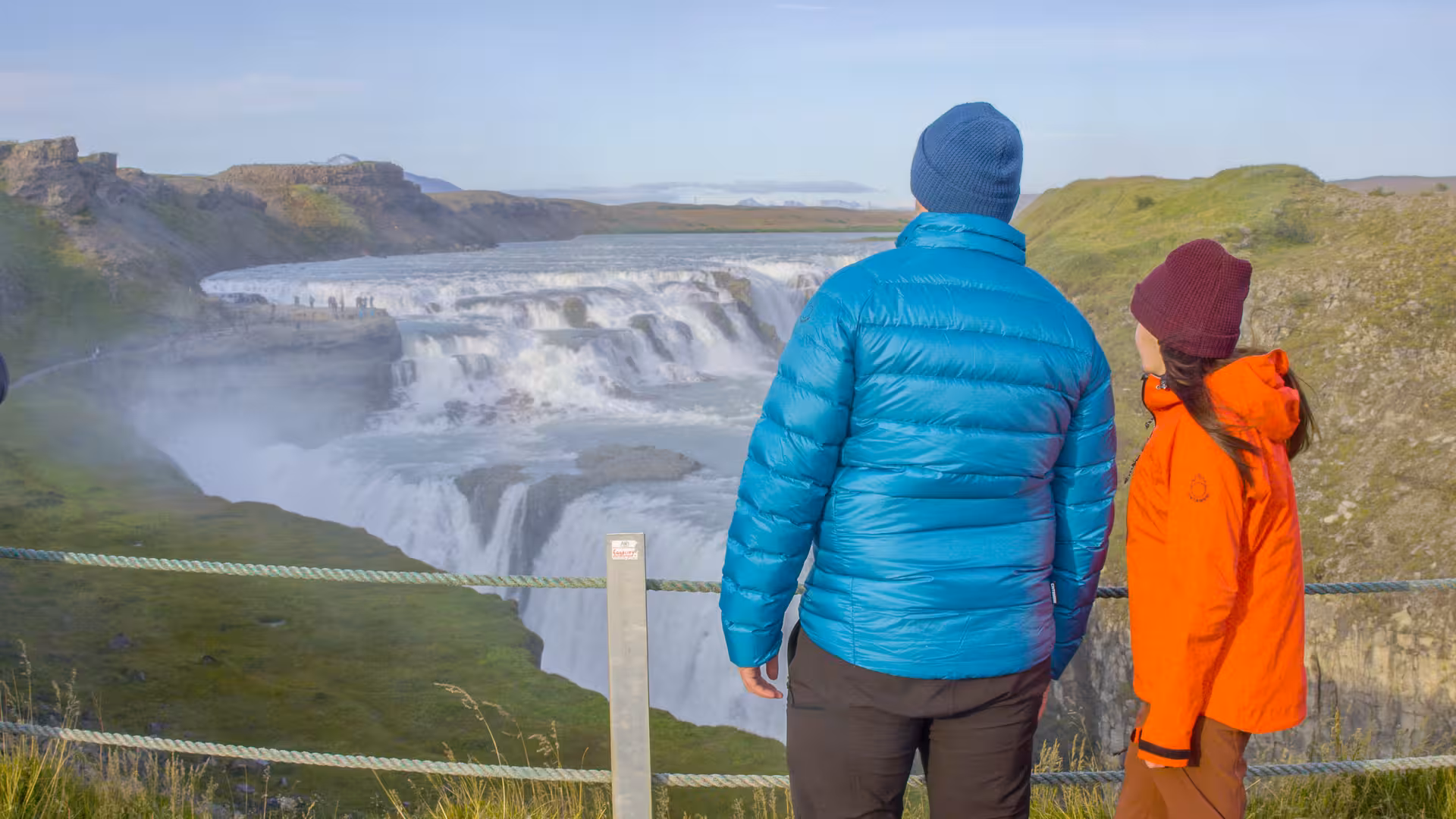 Two tourists in colorful jackets admire the majestic Gullfoss waterfall on a clear day in Iceland's Golden Circle.