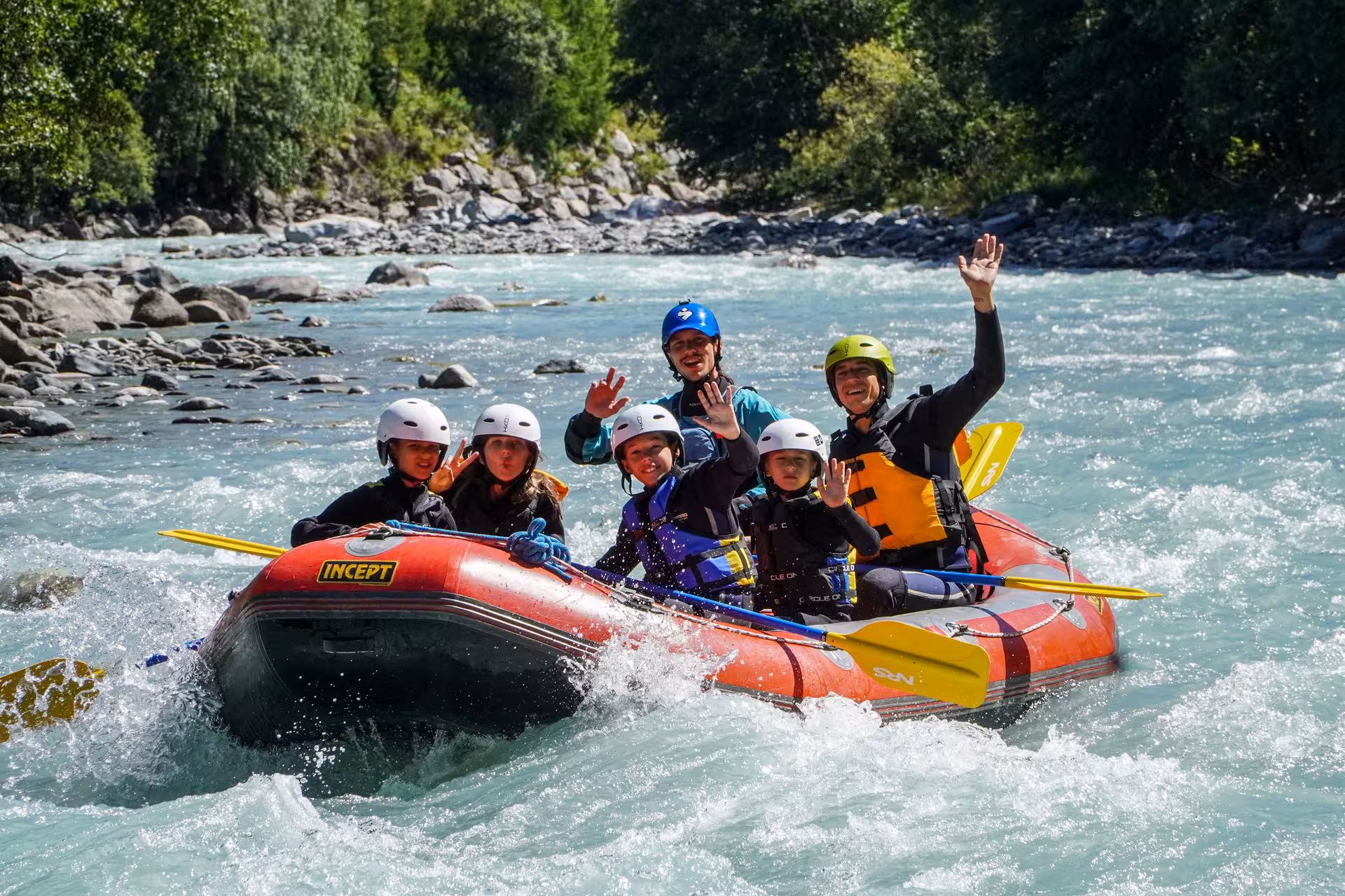 Happy family waving from a raft on a beautiful turquoise river, enjoying Fun for All Rafting in Engadin.