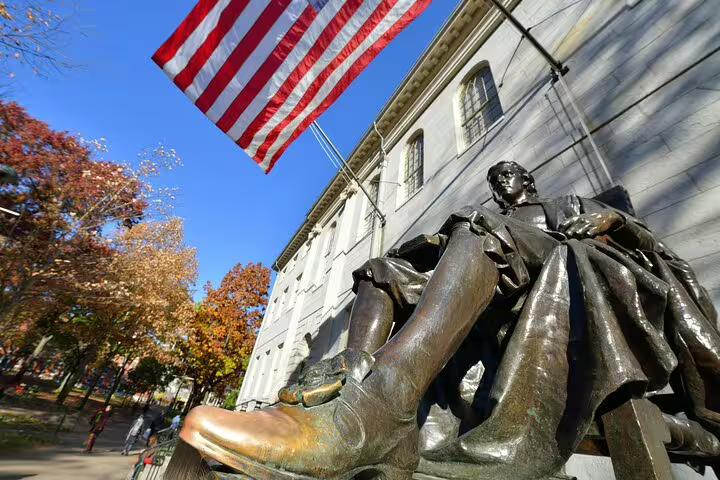Bronze Harvard statue with worn golden boot and American flag outside University Hall in autumn