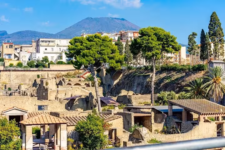 Vibrant view of Herculaneum ruins with lush greenery and Mount Vesuvius in the background on a sunny day.