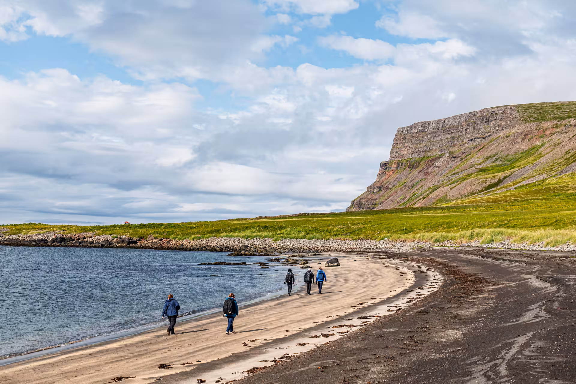 Hikers walking a black-sand beach by cliffs in Hesteyri, Westfjords, on a heritage and nature tour