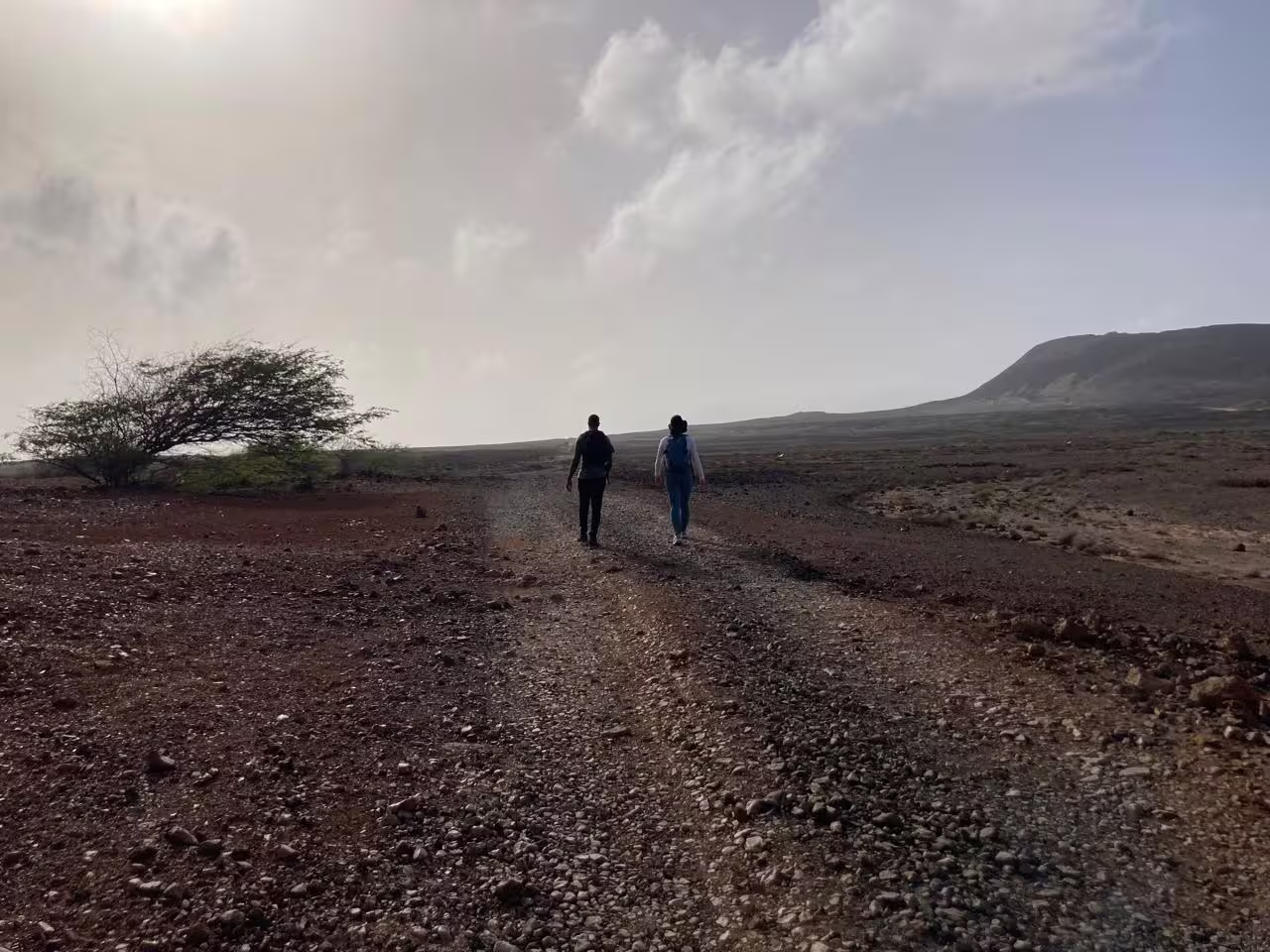 Two hikers venture across a rocky path under a cloudy sky towards Volcano Viana, São Vicente, highlighting adventure tourism.