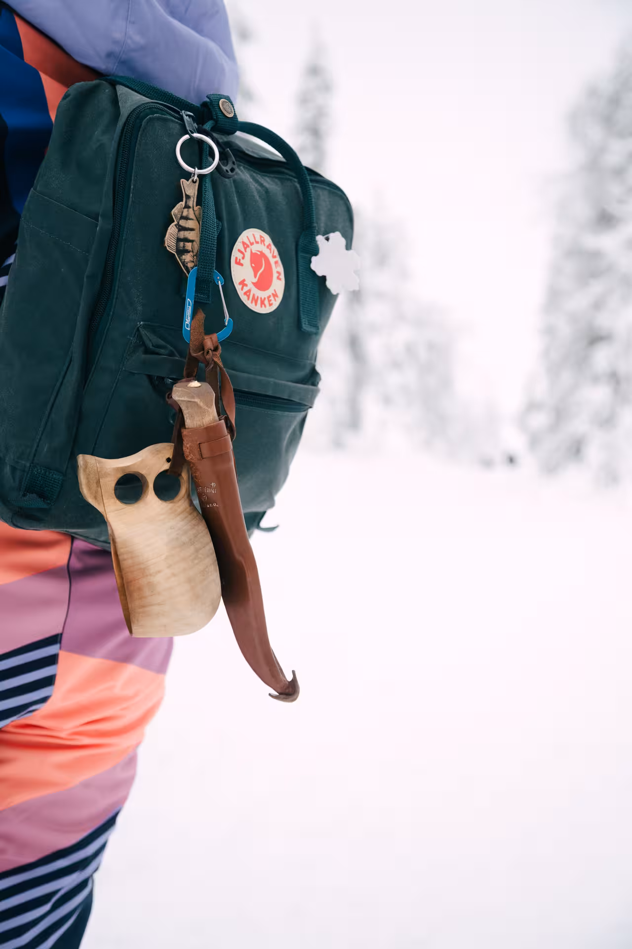 Close-up of a hiker's colorful backpack with wooden cup and knife in snowy Riisitunturi National Park, Finland.