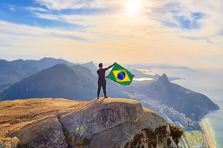 Hiker holding Brazilian flag atop Pedra da Gávea, enjoying stunning panoramic views of Rio de Janeiro's coastline.