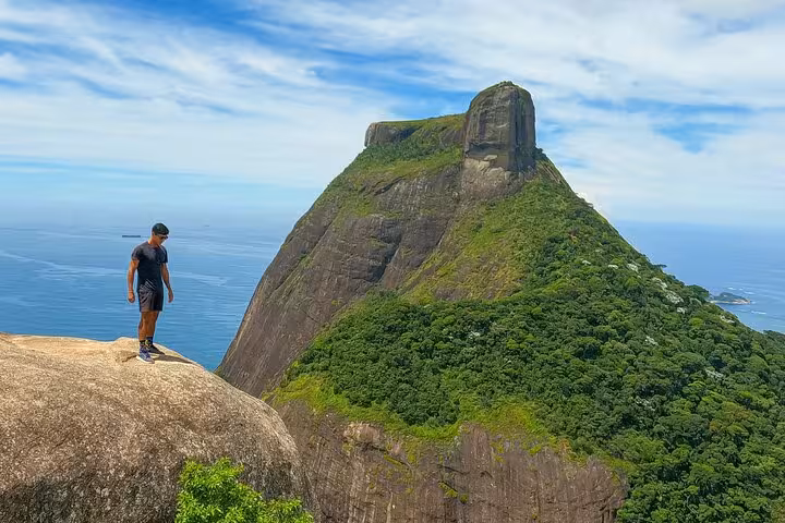 Hiker stands on a rocky ledge with a stunning view of the massive stone peak in Tijuca Forest, Rio de Janeiro.