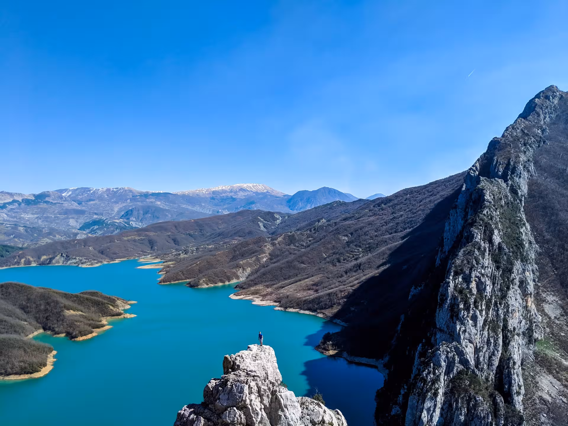 Hiker atop rocky peak overlooking vibrant turquoise Bovilla Lake surrounded by rugged mountains and clear blue skies.